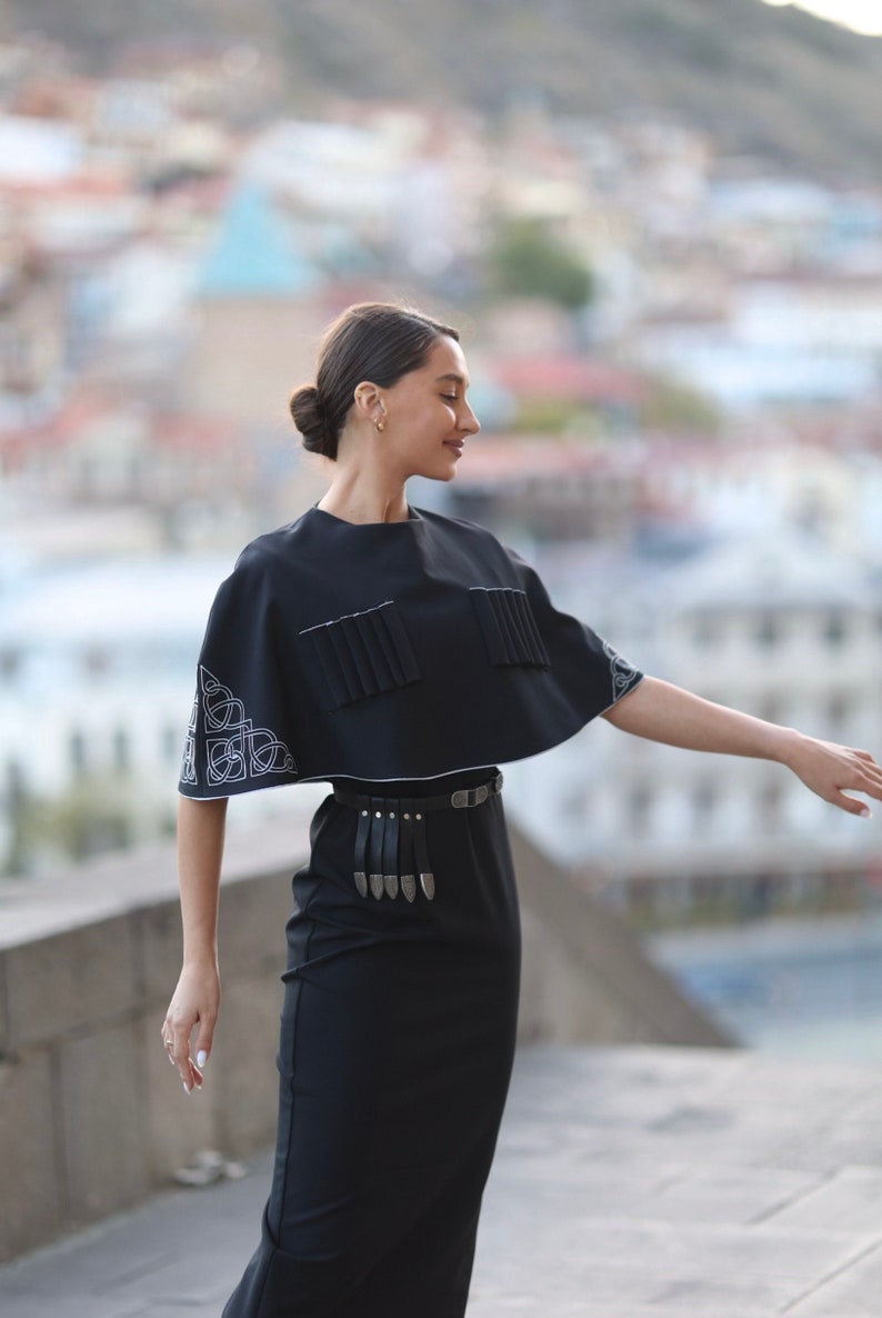 May include: A woman wearing a black dress with a black cape top and a silver belt with a fringe. The cape top has white embroidery on the sleeves.