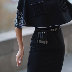 May include: A black dress with a short, cape-like top featuring white embroidered designs. The dress has a black leather belt with silver accents.