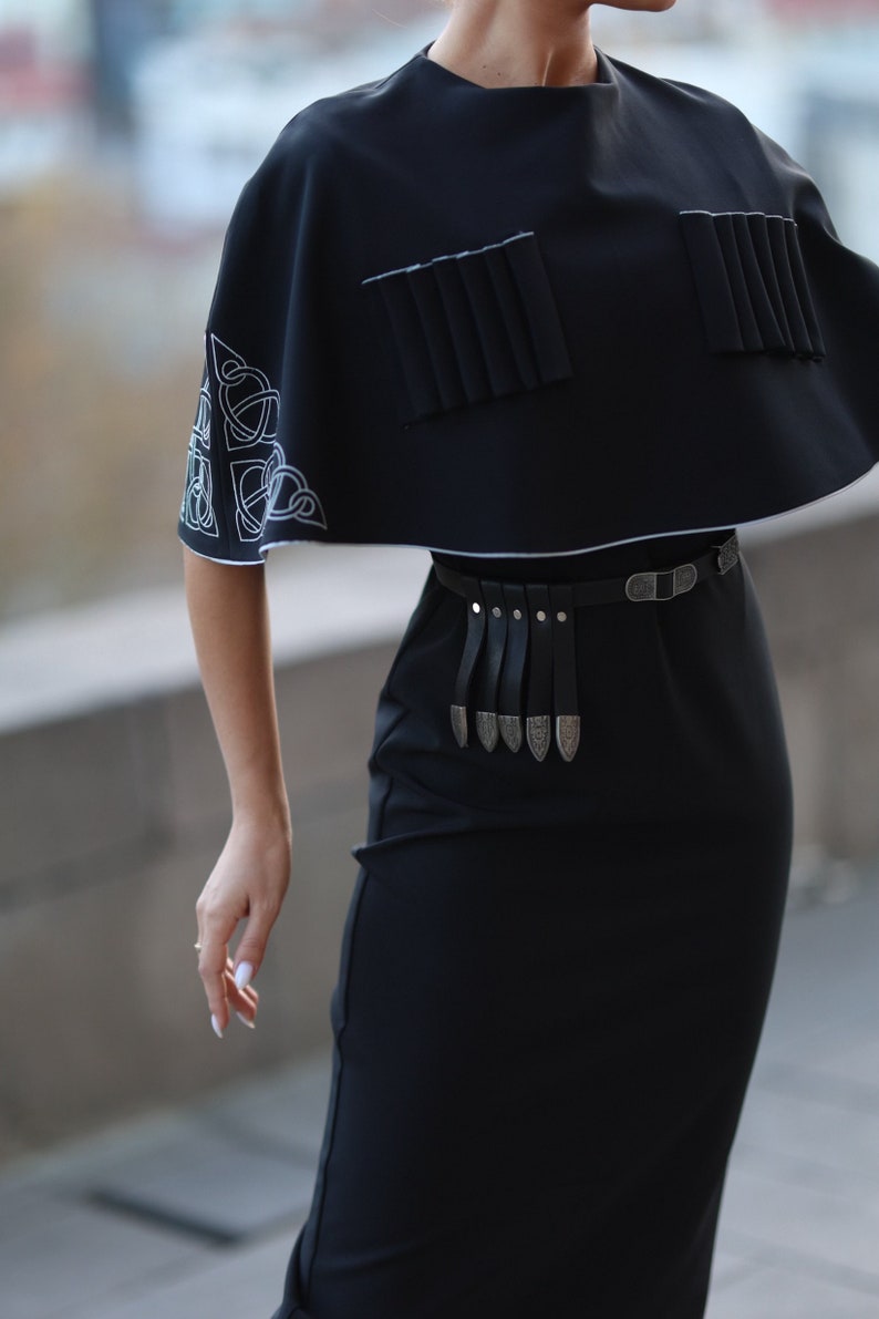 May include: A black dress with a short, cape-like top featuring white embroidered designs. The dress has a black leather belt with silver accents.