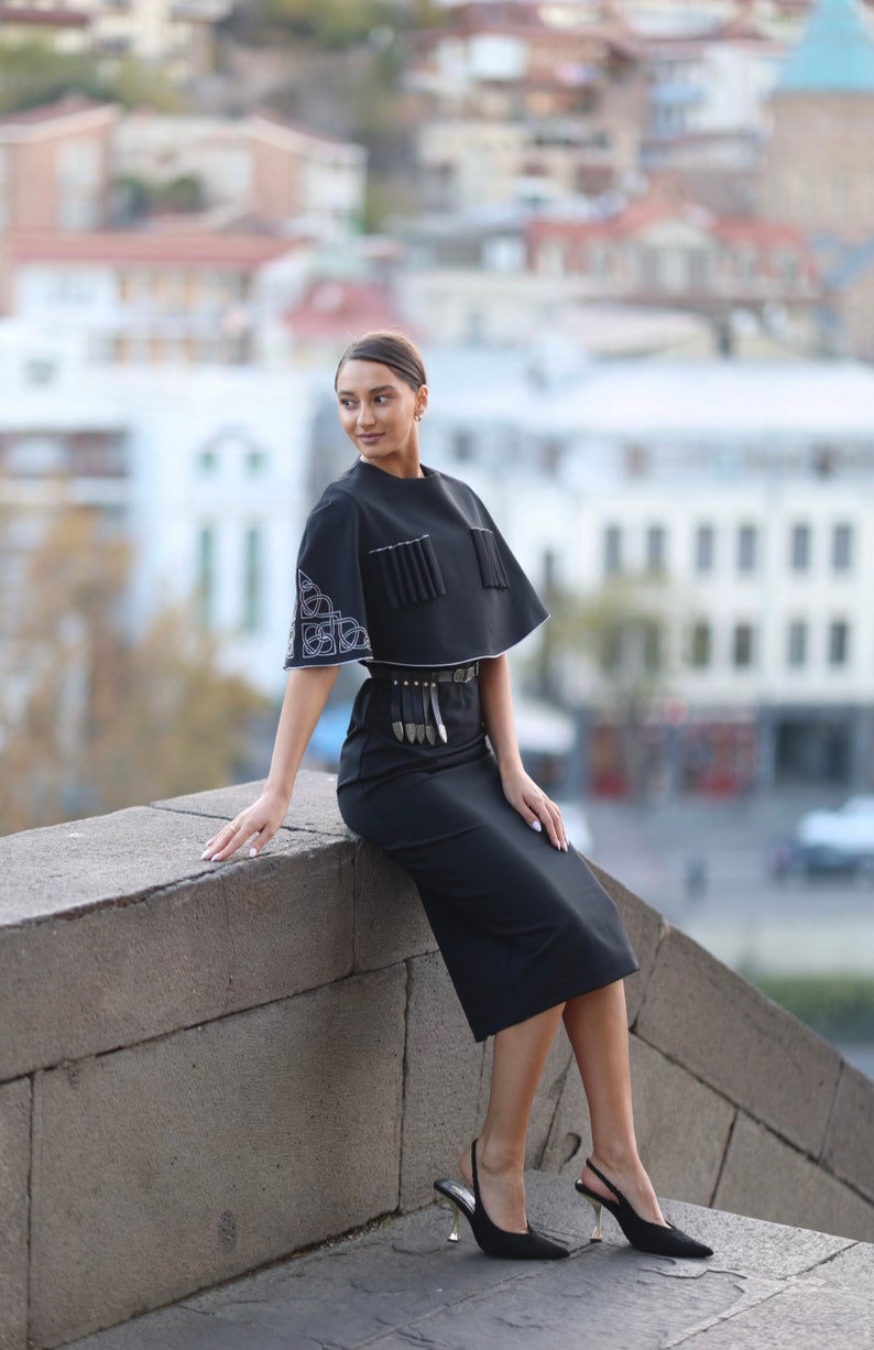 May include: A woman wearing a black skirt and a black top with white embroidery. She is sitting on a stone wall with a city in the background.