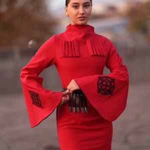 Puede incluir: Un vestido rojo con cuello alto y mangas largas. Las mangas tienen bordados negros y el vestido tiene un cintur&oacute;n negro con detalles plateados.