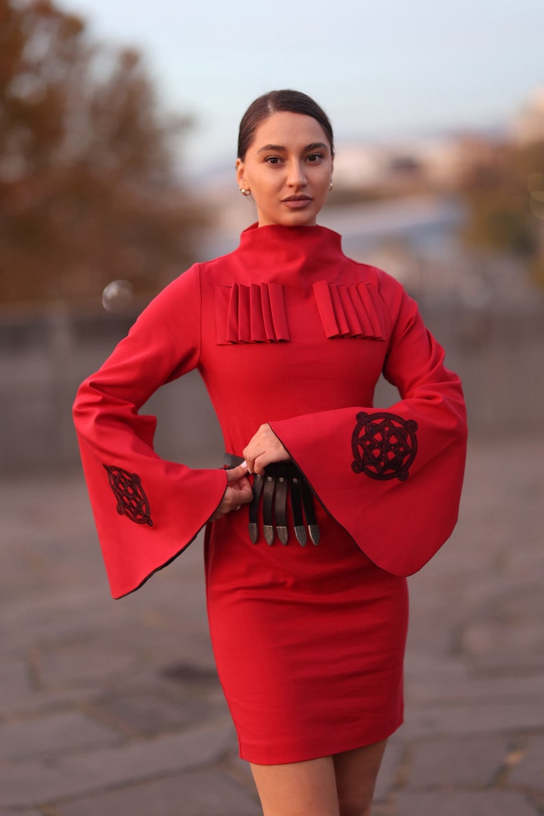 Puede incluir: Un vestido rojo con cuello alto y mangas largas. Las mangas tienen bordados negros y el vestido tiene un cintur&oacute;n negro con detalles plateados.