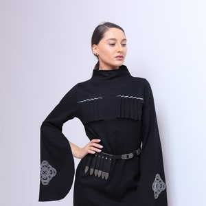 Puede incluir: Un vestido negro de manga larga y cuello alto. El vestido tiene un ribete blanco alrededor del cuello y un cintur&oacute;n plateado con una hebilla decorativa. Las mangas tienen un ribete blanco y un dise&ntilde;o bordado plateado.