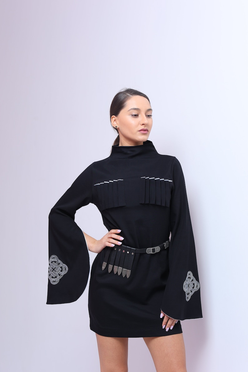 Puede incluir: Un vestido negro de manga larga y cuello alto. El vestido tiene un ribete blanco alrededor del cuello y un cintur&oacute;n plateado con una hebilla decorativa. Las mangas tienen un ribete blanco y un dise&ntilde;o bordado plateado.