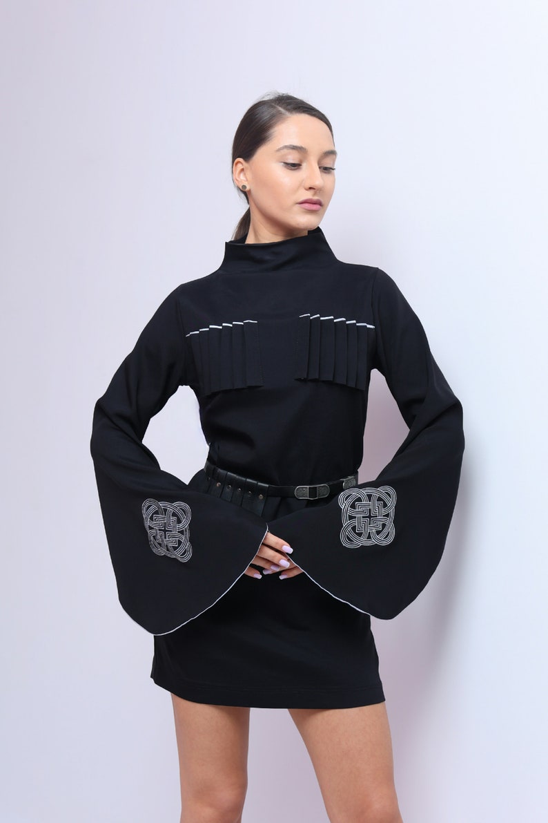Puede incluir: Un vestido negro con mangas largas y un ribete blanco. Las mangas tienen un dise&ntilde;o bordado blanco. El vestido tiene un cintur&oacute;n negro.