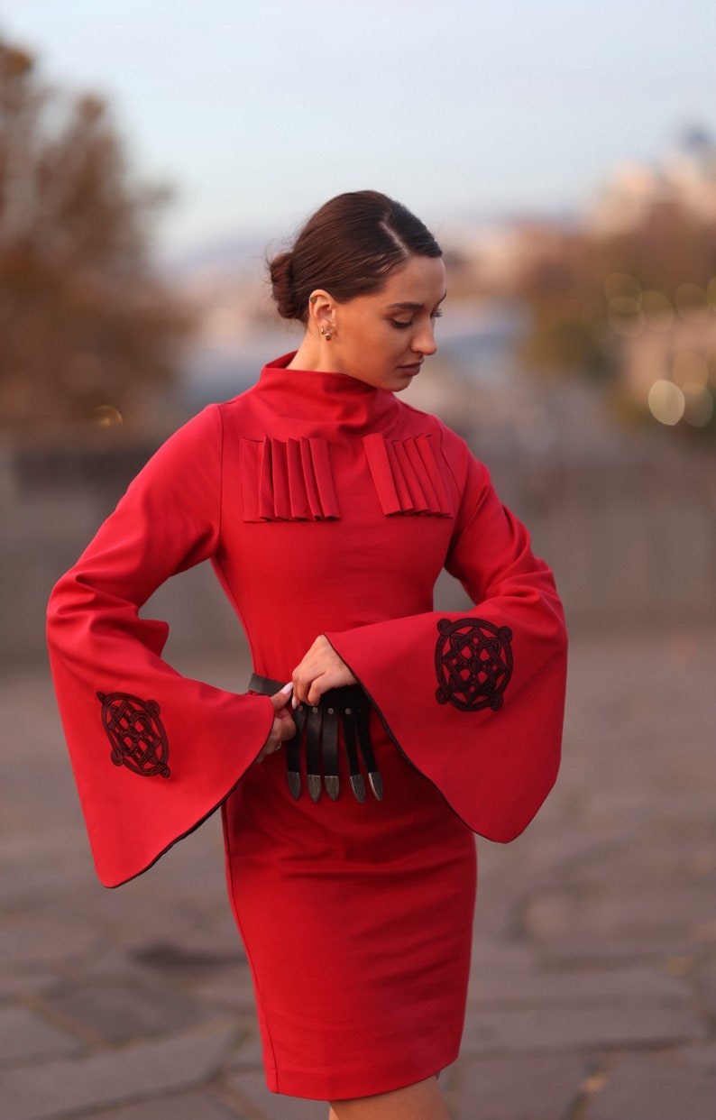 Puede incluir: Un vestido rojo con mangas largas y un cintur&oacute;n negro. Las mangas tienen bordados negros con un dise&ntilde;o circular. El vestido tiene un cuello alto y una silueta ajustada.
