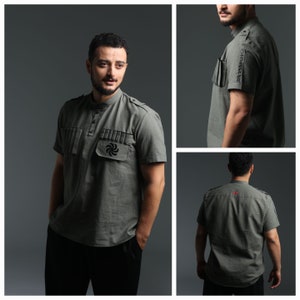 Könnte beinhalten: Ein grünes, kurzärmeliges Hemd mit Stehkragen und einer kleinen Tasche auf der Brust. Die Tasche hat ein schwarzes gesticktes Design. Das Hemd hat einen Knopfverschluss am Hals.