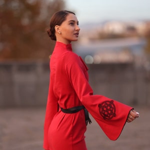 Puede incluir: Una mujer con un vestido rojo con cintur&oacute;n negro y mangas bordadas. El vestido tiene un cuello alto y mangas largas.