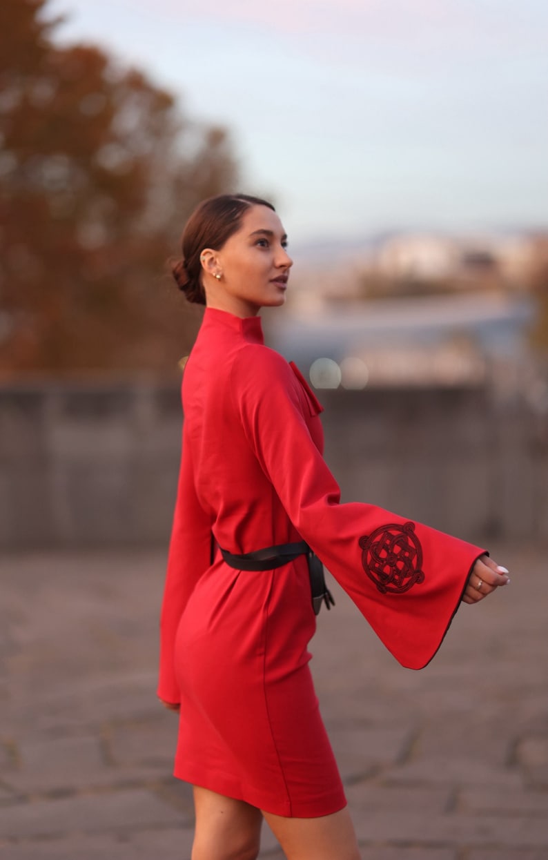 Puede incluir: Una mujer con un vestido rojo con cintur&oacute;n negro y mangas bordadas. El vestido tiene un cuello alto y mangas largas.