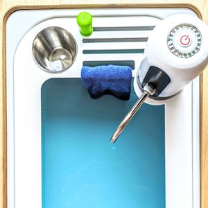 May include: A white countertop sink with a blue basin, a water dispenser with a silver spout, a green knob, and a power button. A blue dishcloth hangs on the side of the sink.