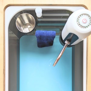 May include: A white water dispenser with a blue and gray base, a copper spout, and a power button. The dispenser is in a gray sink with a blue basin. A blue cloth and a silver cup are in the sink.
