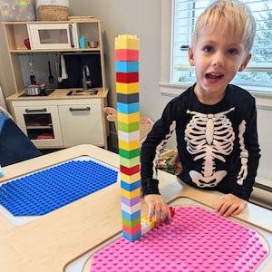 May include: A child builds a tower with colourful Lego blocks on a wooden table with pink and blue Lego baseplates.