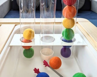 May include: A sensory activity set with clear tubes holding colorful felt balls in a rainbow spectrum. Additional felt balls and tongs are in a white tray. Rainbow decor is visible in the background.