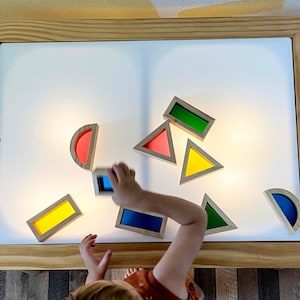 May include: A child is playing with colourful geometric shapes on a light table. The shapes are made of wood and have translucent coloured glass inserts. The table has a white light panel and a wooden frame.