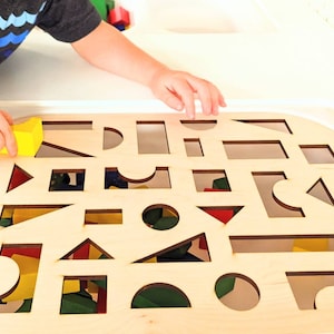 May include: A wooden board with cut-out shapes for a child's shape sorter toy. The board is light brown and has a variety of shapes cut out, including circles, squares, triangles, and rectangles. A child's hand is shown placing a yellow block into one of the cut-out shapes.