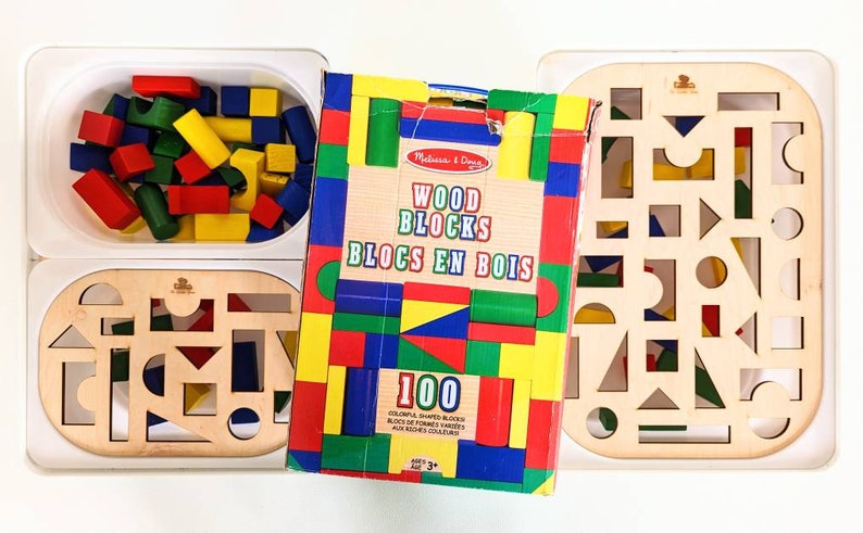 May include: A box of 100 colorful wooden blocks with the text "Wood Blocks" and "Blocs en Bois" on the front. The box is open and shows the blocks inside. There are also two wooden trays with cutouts for the blocks.