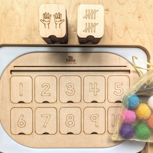 May include: Wooden counting toy with a tray featuring numbers 1-10. The tray has a slot for placing counting tokens. There are two stacks of wooden blocks, one with hand symbols and the other with tally marks. A small bag of colorful felt balls is included.