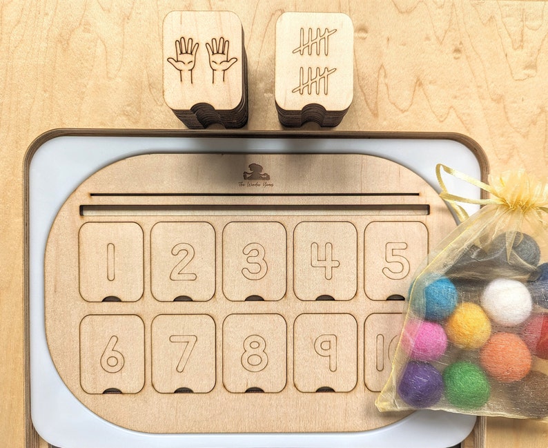 May include: Wooden counting toy with a tray featuring numbers 1-10. The tray has a slot for placing counting tokens. There are two stacks of wooden blocks, one with hand symbols and the other with tally marks. A small bag of colorful felt balls is included.