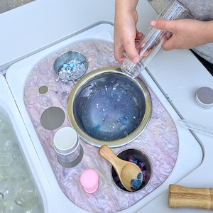 May include: A white plastic sensory table with a variety of compartments for different activities. The table has a pink and white marbled surface. A child's hand is pouring blue and white glitter into a clear plastic container. The container is being poured into a silver bowl filled with water. There is a wooden spoon, a pink rubber duck, and a small silver bowl filled with blue and purple gems.