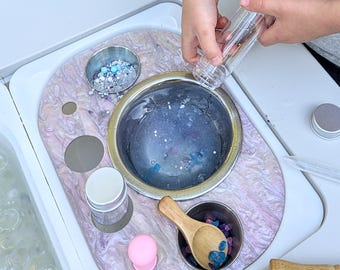 May include: A sensory play station with a purple and white marbled surface. It features a large stainless steel bowl filled with blue glitter and small containers with various sensory materials, including a wooden scoop and a clear tube.