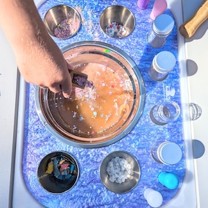 May include: A blue and purple tray with eight stainless steel bowls, four clear glass bottles with silver lids, two pink bottles with silver lids, a blue bottle with a silver lid, a white bottle with a silver lid, a wooden spoon, and a wooden mallet. The bowls contain various colorful sprinkles, glitter, and white sugar.