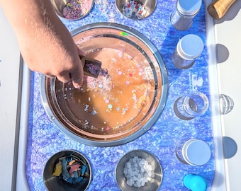 May include: A sensory play station featuring a blue marbled surface with a large central stainless steel bowl containing liquid and colorful sprinkles. Four smaller stainless steel bowls hold various materials, alongside several small jars and lids.