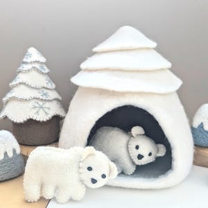Może przedstawiać: Dwa białe filcowe niedźwiedzie polarne, jeden wewnątrz białego filcowego igloo, a drugi na zewnątrz. Igloo ma czarny filcowy otwór i warstwowy biały filcowy dach. Mała niebieska filcowa góra znajduje się w tle.