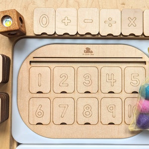 May include: Wooden math manipulative set with number tiles, operation symbols, and a frame with number slots. The frame has the numbers 1 through 10. There are also colorful felt balls in a mesh bag.