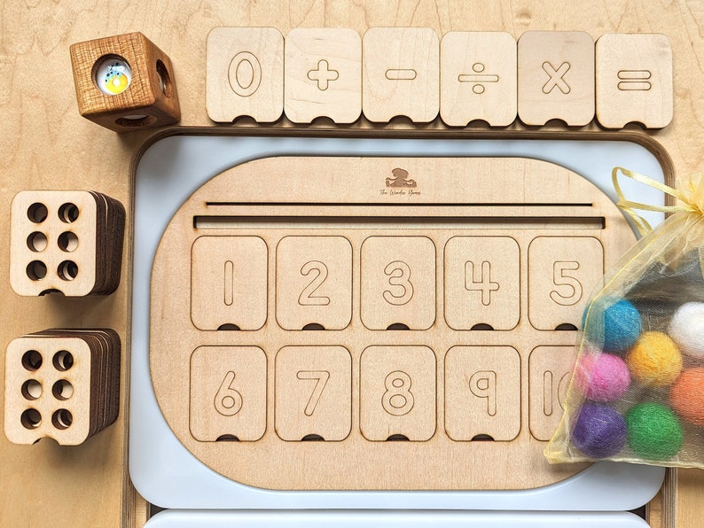 May include: Wooden math manipulative set with number tiles, operation symbols, and a frame with number slots. The frame has the numbers 1 through 10. There are also colorful felt balls in a mesh bag.