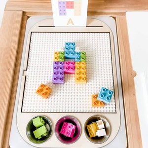 Könnte beinhalten: Eine weiße Stecktafel mit einer Karte mit dem Buchstaben A und einem bunten Blockpuzzle. Das Puzzle besteht aus Blöcken in verschiedenen Farben, darunter Blau, Grün, Gelb, Rosa und Lila. Die Blöcke sind in Form des Buchstabens A angeordnet. Es gibt drei kleine Schalen mit zusätzlichen Blöcken.
