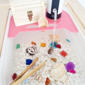 K&ouml;nnte beinhalten: Ein sensorischer Wassertisch mit wei&szlig;en Kieselsteinen, bunten Glassteinen und Muscheln. Eine h&ouml;lzerne Schatztruhe, eine kleine Holzschaufel und eine Bambuszange sind enthalten. Eine wei&szlig;-schwarze elektrische Wasserpumpe gibt Wasser ab.