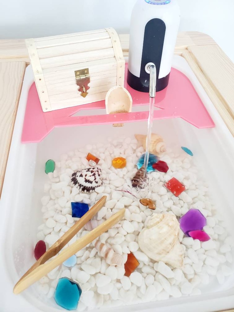 K&ouml;nnte beinhalten: Ein sensorischer Wassertisch mit wei&szlig;en Kieselsteinen, bunten Glassteinen und Muscheln. Eine h&ouml;lzerne Schatztruhe, eine kleine Holzschaufel und eine Bambuszange sind enthalten. Eine wei&szlig;-schwarze elektrische Wasserpumpe gibt Wasser ab.