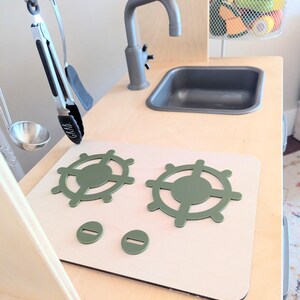 Pu&ograve; includere: Una cucina giocattolo in legno bianco con un lavello grigio, un piano cottura verde con due fornelli e un portautensili in metallo.