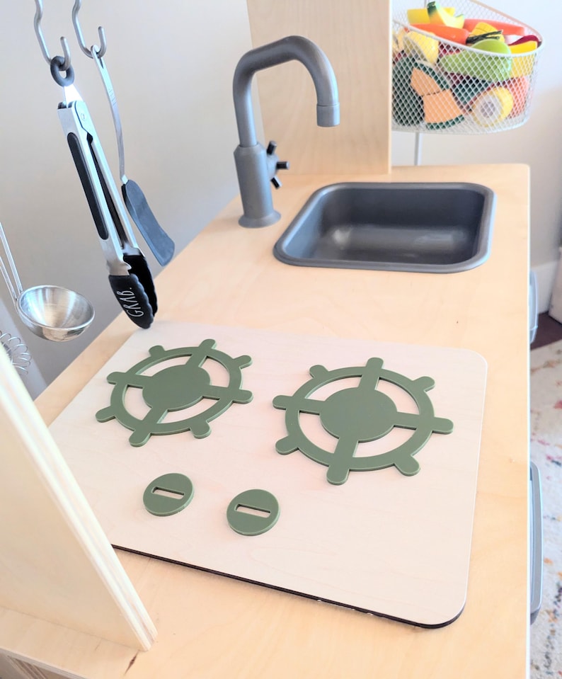 Pu&ograve; includere: Una cucina giocattolo in legno bianco con un lavello grigio, un piano cottura verde con due fornelli e un portautensili in metallo.