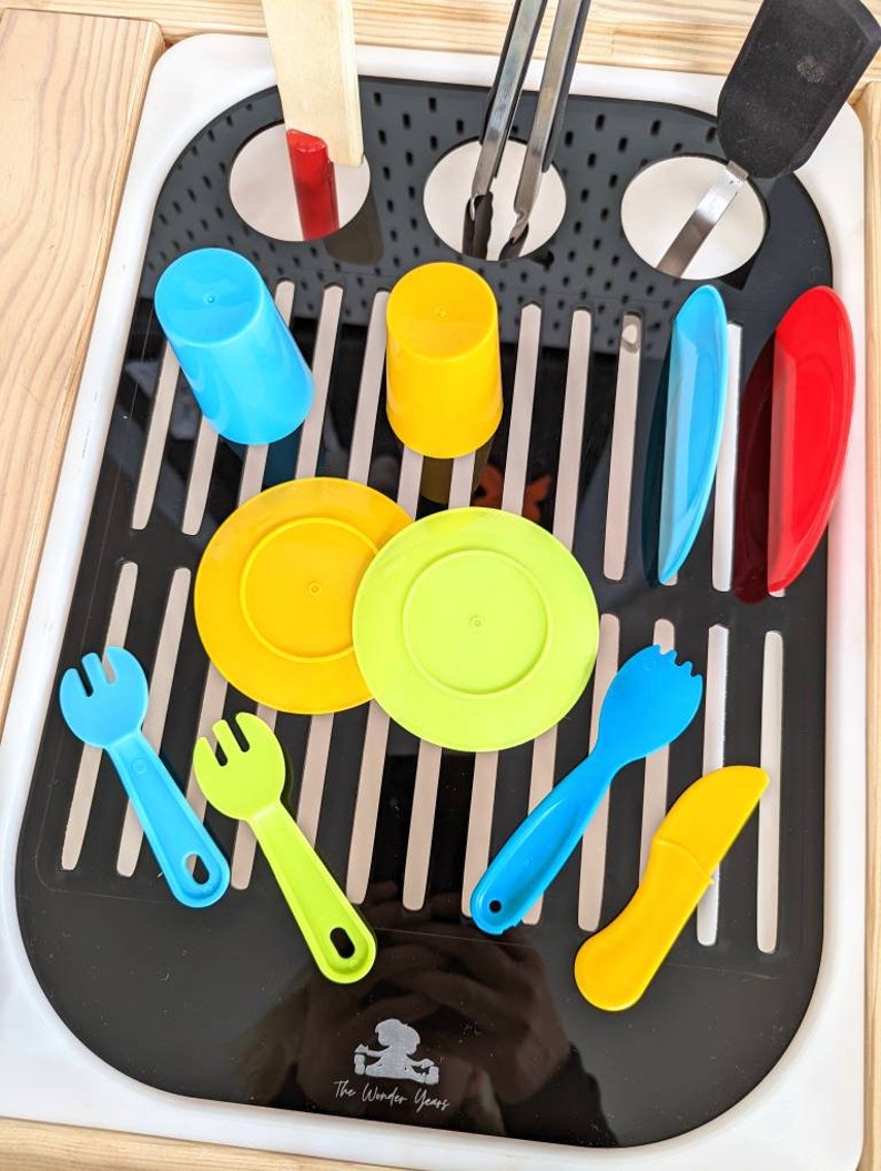 Puede incluir: Un escurridor de platos de pl&aacute;stico negro con varios art&iacute;culos de cocina de pl&aacute;stico de colores, incluyendo tazas, platos, tenedores, un cuchillo y una esp&aacute;tula. El escurridor tiene un borde blanco y est&aacute; sobre una superficie de madera. El texto "The Wonder Years" es visible en la parte inferior del escurridor.