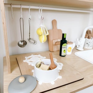Pu&ograve; includere: Un bancone da cucina giocattolo bianco con un tagliere di legno, una pentola bianca con coperchio, un cucchiaio di legno e un piccolo sacchetto di limoni gialli. Il bancone ha una griglia di metallo bianca con ganci per appendere gli utensili.