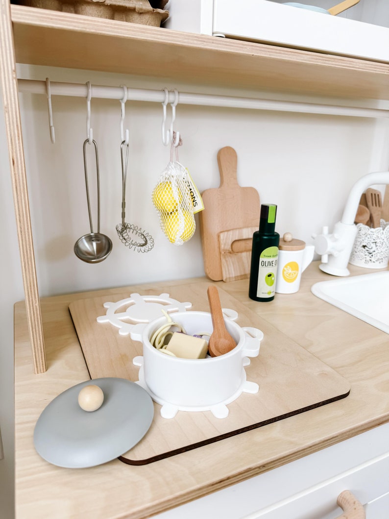 Pu&ograve; includere: Un bancone da cucina giocattolo bianco con un tagliere di legno, una pentola bianca con coperchio, un cucchiaio di legno e un piccolo sacchetto di limoni gialli. Il bancone ha una griglia di metallo bianca con ganci per appendere gli utensili.