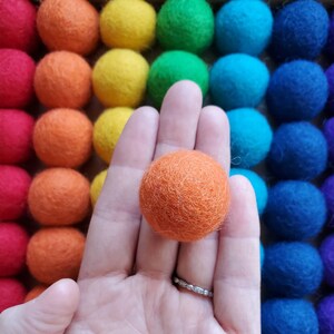 May include: A hand holding a single orange felt ball against a background of colorful felt balls.