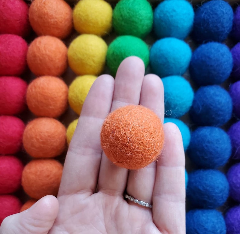 May include: A hand holding a single orange felt ball against a background of colorful felt balls.