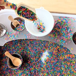 May include: A white plastic sensory bin filled with colorful rice. There are four small metal cups with wooden spoons, a white bowl, and a wooden scoop filled with rice.