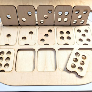 May include: A wooden game board with nine compartments, each containing a different number of holes. The board is made of light wood and has a rounded edge. There are also five separate wooden pieces with different numbers of holes.