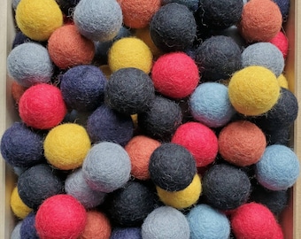 May include: A wooden box filled with an assortment of colorful felt balls. The balls are in various shades of blue, yellow, orange, red, and gray. The felt balls are densely packed together, creating a textured and vibrant display.