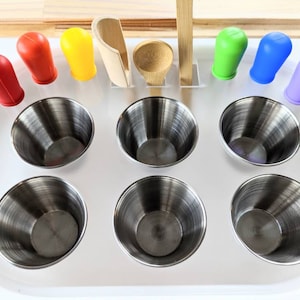 May include: A white tray with eight stainless steel cups and seven colourful silicone finger puppets. The tray is designed for sensory play and includes two wooden spoons and a wooden scoop.