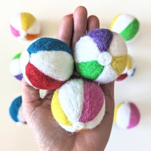Peut inclure: Une main tenant une variété de balles de plage en feutre de différentes couleurs. Les balles sont de couleurs et de motifs différents, notamment rouge, blanc, bleu, vert, jaune et violet.