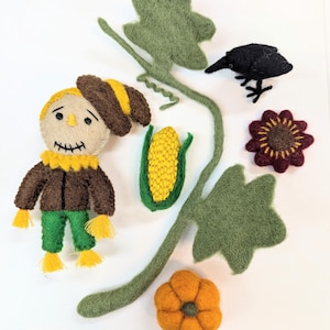 May include: Felt craft set featuring a scarecrow, a crow, a pumpkin, a corn cob, and a sunflower. The set is made of felt and has a rustic, autumnal theme.