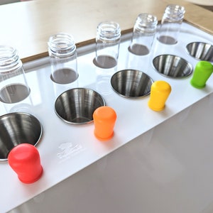 May include: A white plastic tray with six clear glass bottles, six stainless steel funnels, and six colourful silicone stoppers. The stoppers are red, orange, yellow, green, blue, and purple.