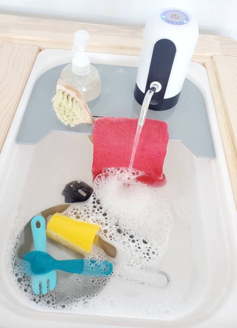 K&ouml;nnte beinhalten: Ein wei&szlig;-schwarzer elektrischer Wasserspender steht auf einer grauen Ablage &uuml;ber einem wei&szlig;en Waschbecken. Ein rotes Tuch, eine Holzb&uuml;rste, eine transparente Seifenflasche, ein gelber Schwamm und blaues Plastikbesteck liegen im Seifenwasser.