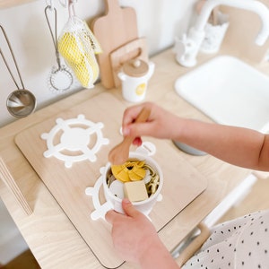 Pu&ograve; includere: Un bambino gioca con un set da cucina giocattolo, mescolando una ciotola di pasta finta con un cucchiaio di legno. La ciotola &egrave; su una superficie di legno con forme di volante in plastica bianca.