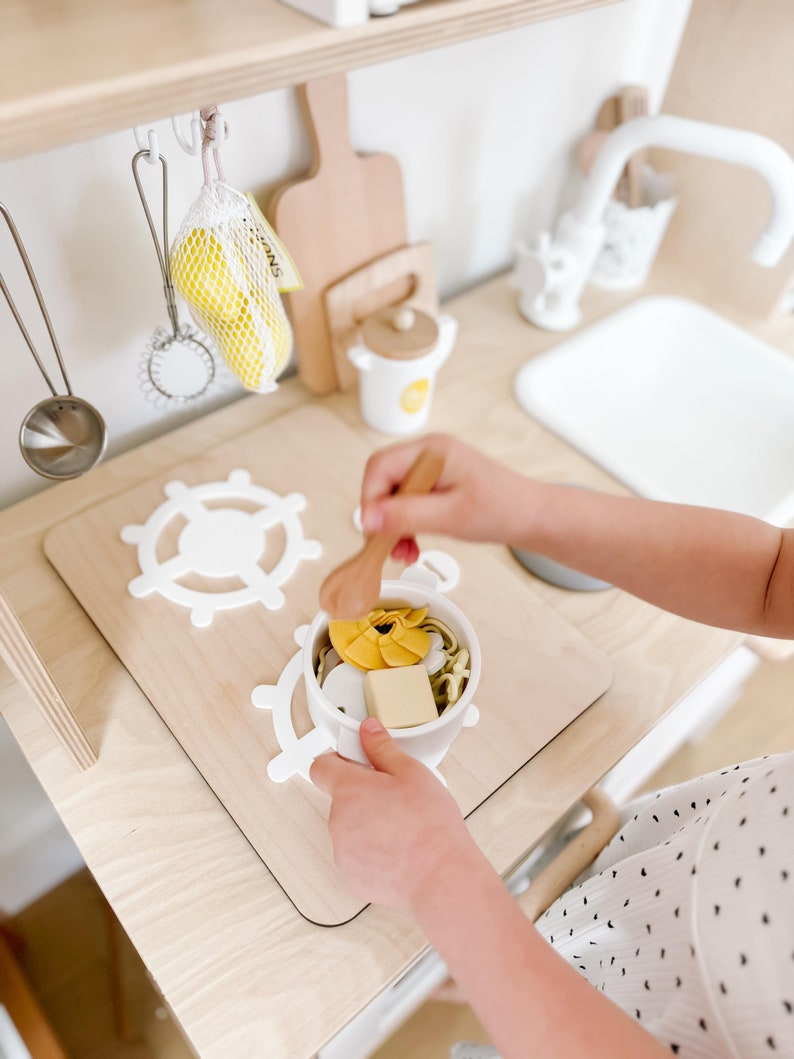 Pu&ograve; includere: Un bambino gioca con un set da cucina giocattolo, mescolando una ciotola di pasta finta con un cucchiaio di legno. La ciotola &egrave; su una superficie di legno con forme di volante in plastica bianca.