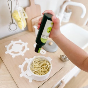 Pu&ograve; includere: Un set da cucina per bambini con un lavello bianco e un piano di lavoro in legno. Una piccola pentola bianca con spaghetti gialli &egrave; sul piano di lavoro. Una bottiglia verde con un'etichetta bianca che dice "110 34" &egrave; tenuta da una mano di bambino. La bottiglia sta venendo versata sugli spaghetti.